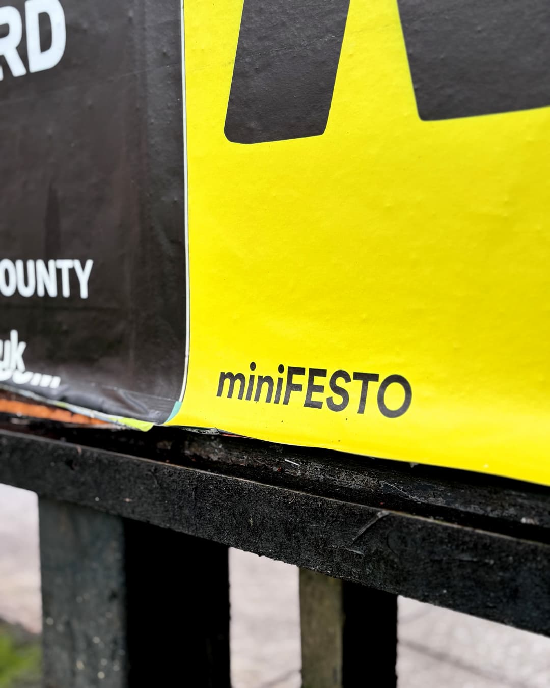 Close-up of a yellow poster with the word "miniFESTO" printed on it, displayed on a black rail.