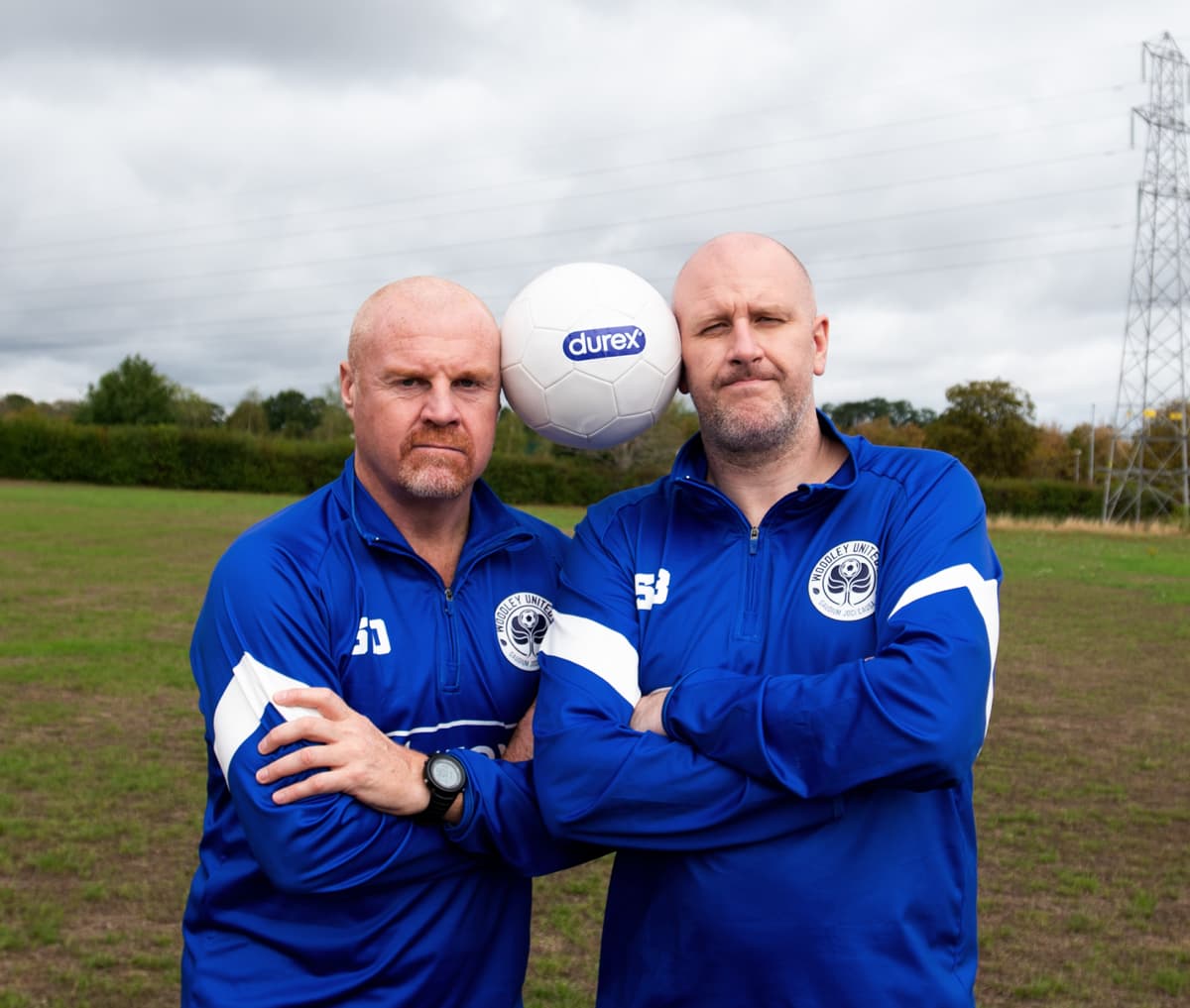 Two people in blue track jackets stand in a field with a football balanced between their heads.