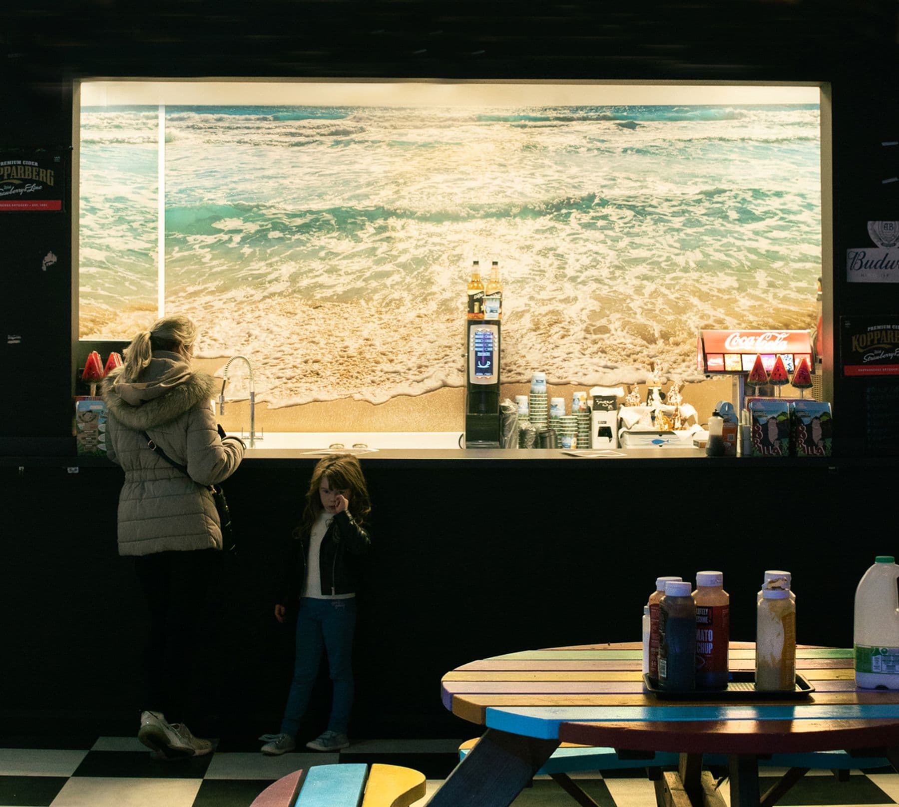 A photograph of a person and a child standing next to a sink at a soft drink dispenser counter. The shot features a table with different sauces and a carton of milk.