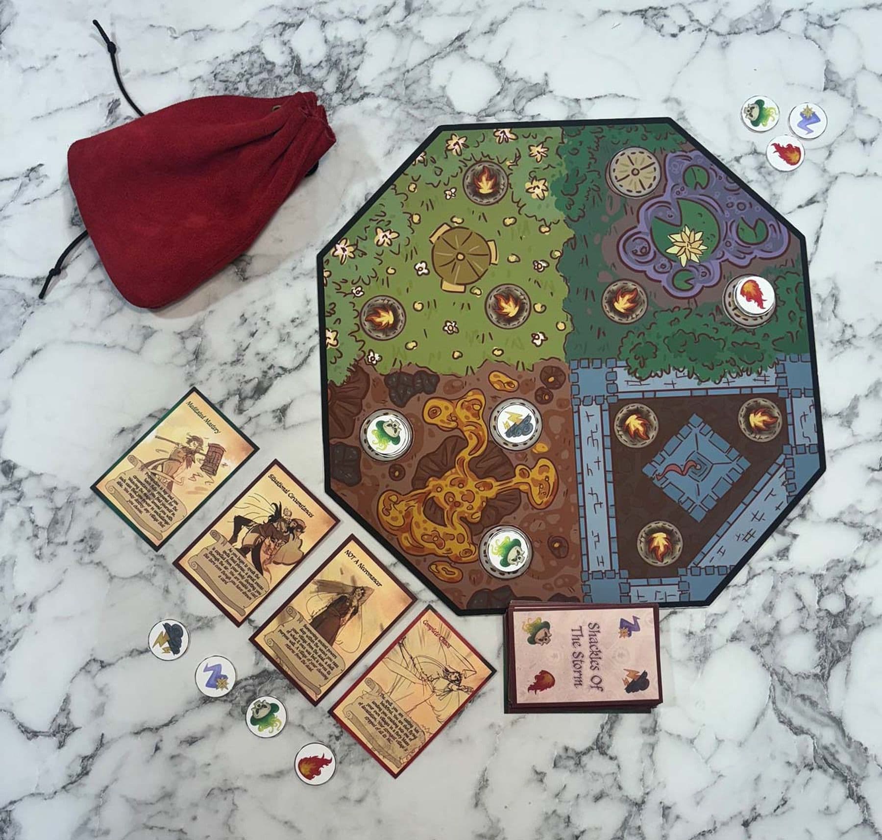 digital mock-up of an octagonal board game on a marble surface, featuring a castle, volcano, forest and field. Counters displaying symbols like fire and lightening are scattered across the board. Nearby, cards with the title Shackles of the Storm are arranged in a pile, with four cards laid next to the pile face up.