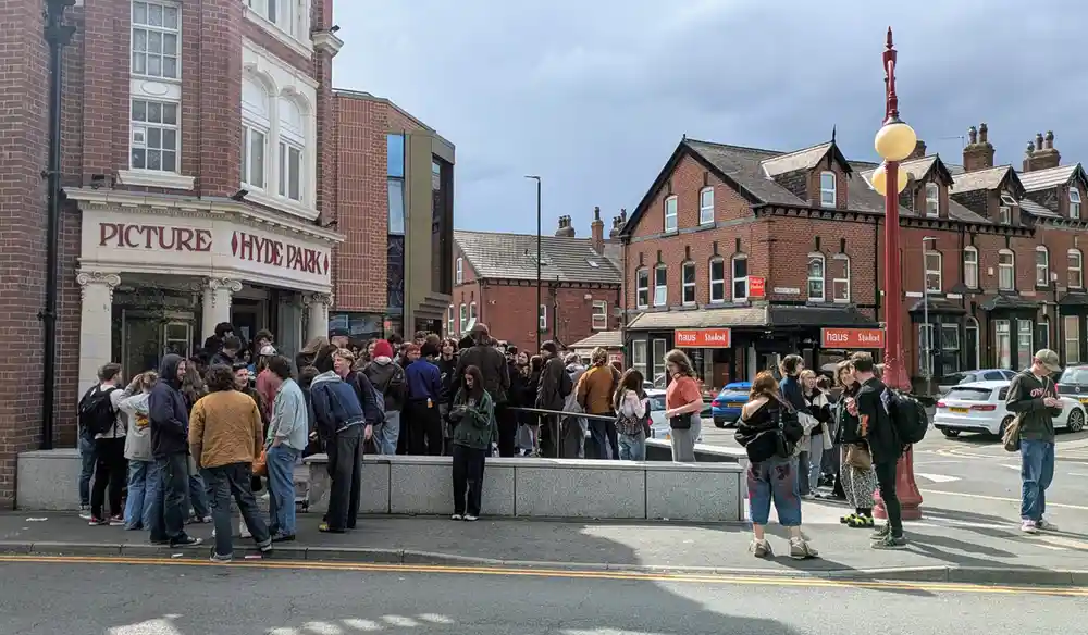 A photograph of a crowd of people gathered outside Hyde Park Picture House, a historic red-brick cinema, with nearby brick buildings and parked cars on a cloudy day.