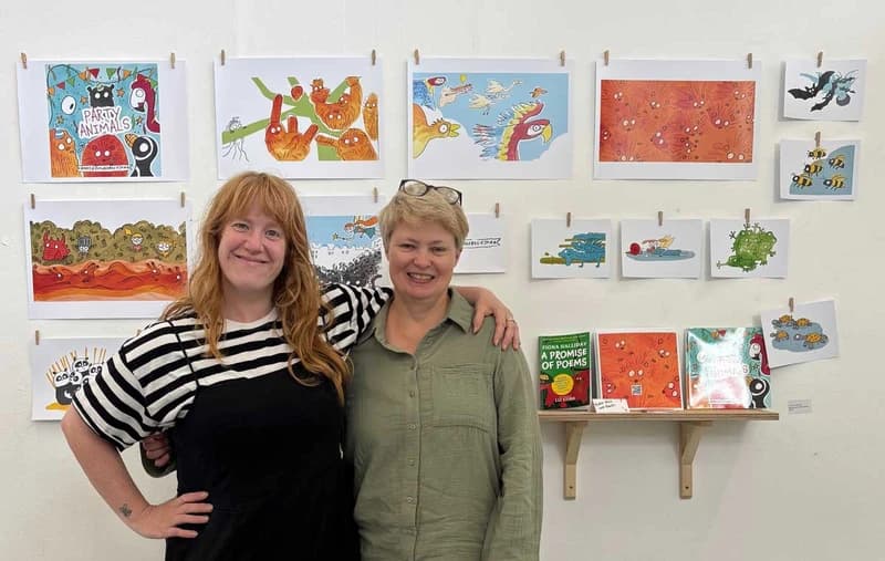 Two smiling people stand arm in arm in front of a wall displaying colorful illustrations and books on a small shelf.