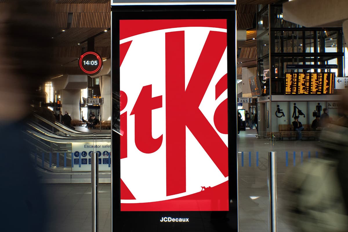 Digital billboard displaying a KitKat ad in a busy train station with blurred commuters and a clock showing 14:05 in the background.