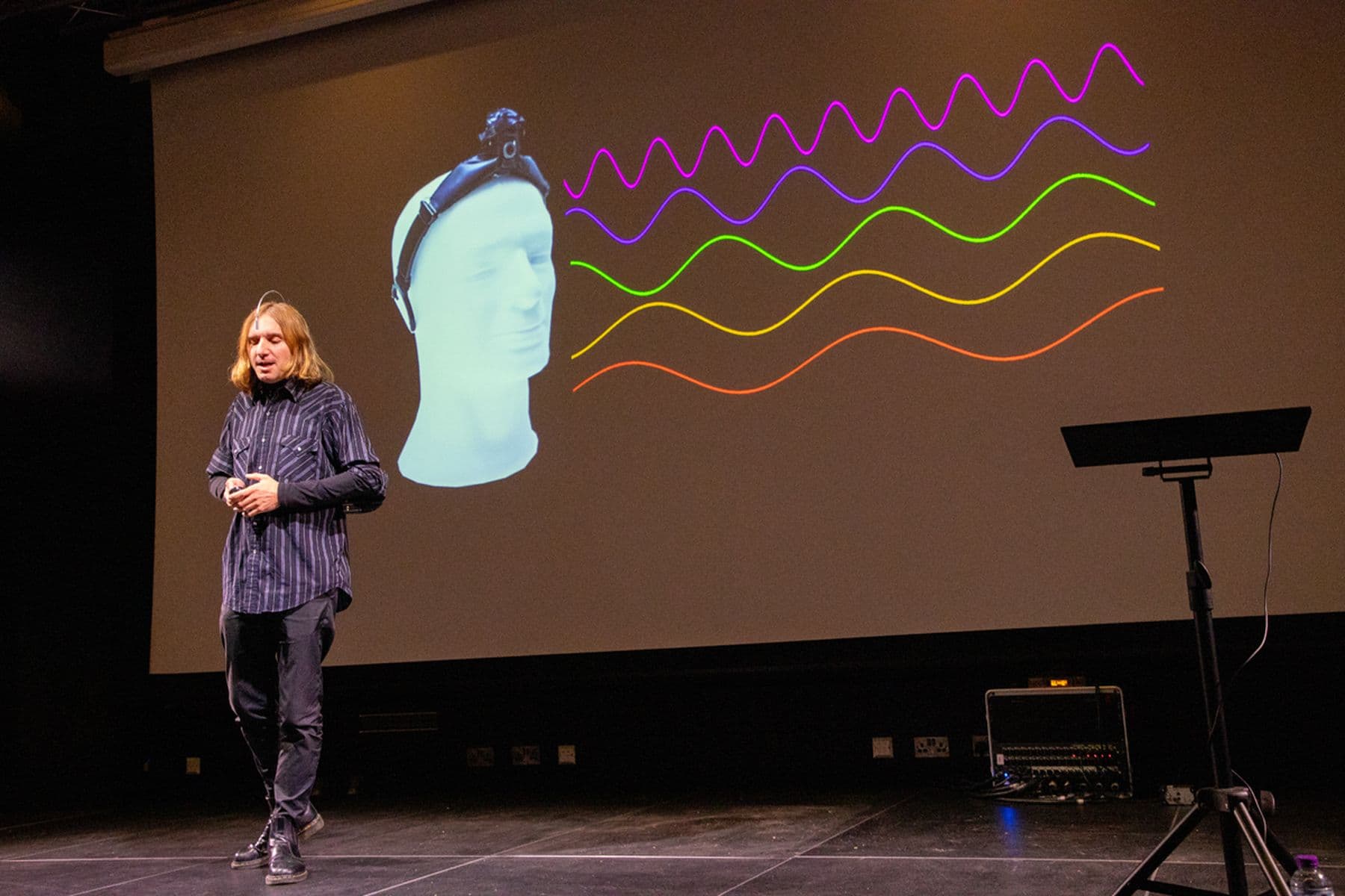 A photograph of a person presenting on stage with a projection showing a mannequin wearing a headband emitting colourful waves on a screen behind.