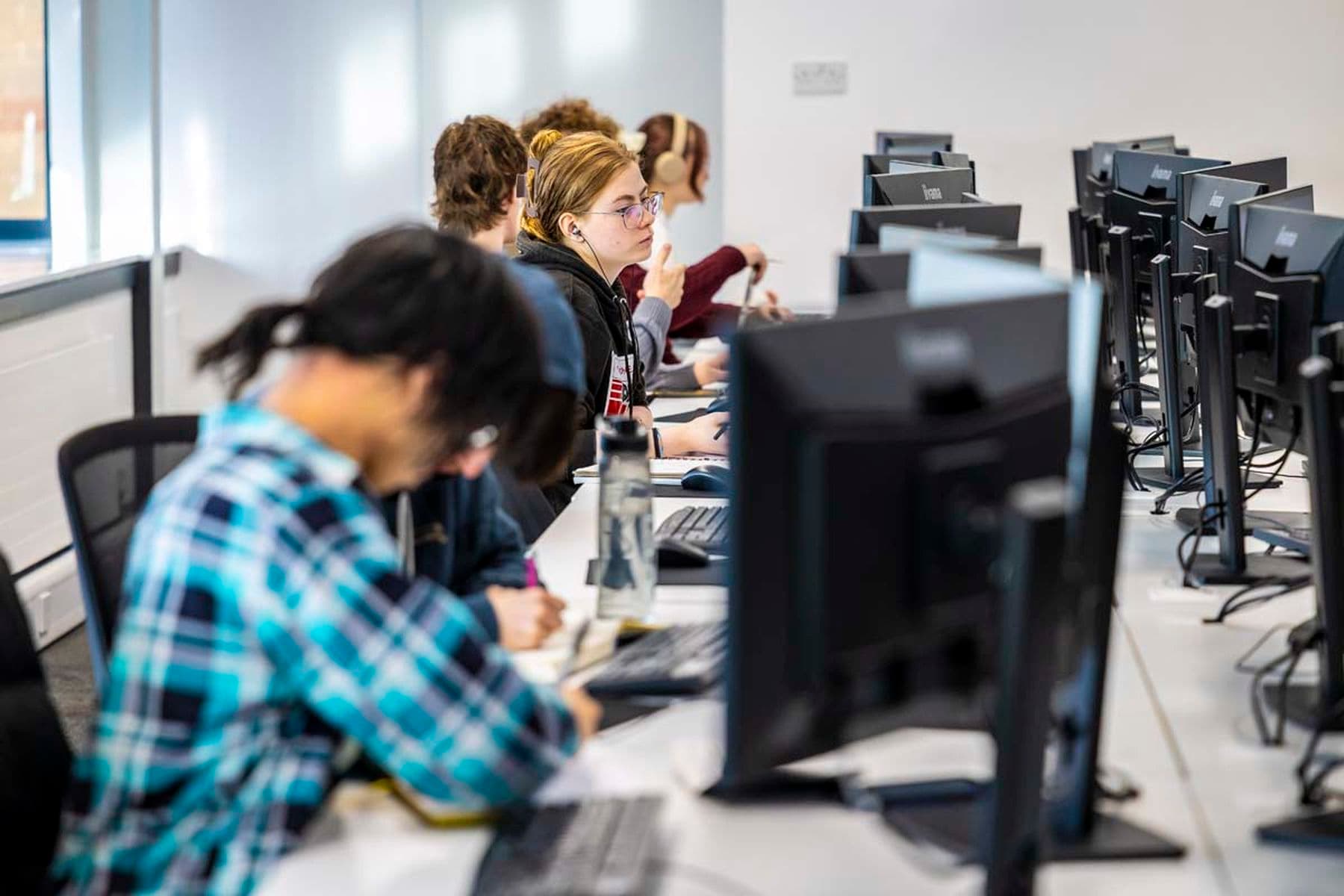 A photograph featuring a row of students sitting at computers, some wearing headphones and others drawing in sketchbooks or on tablets.