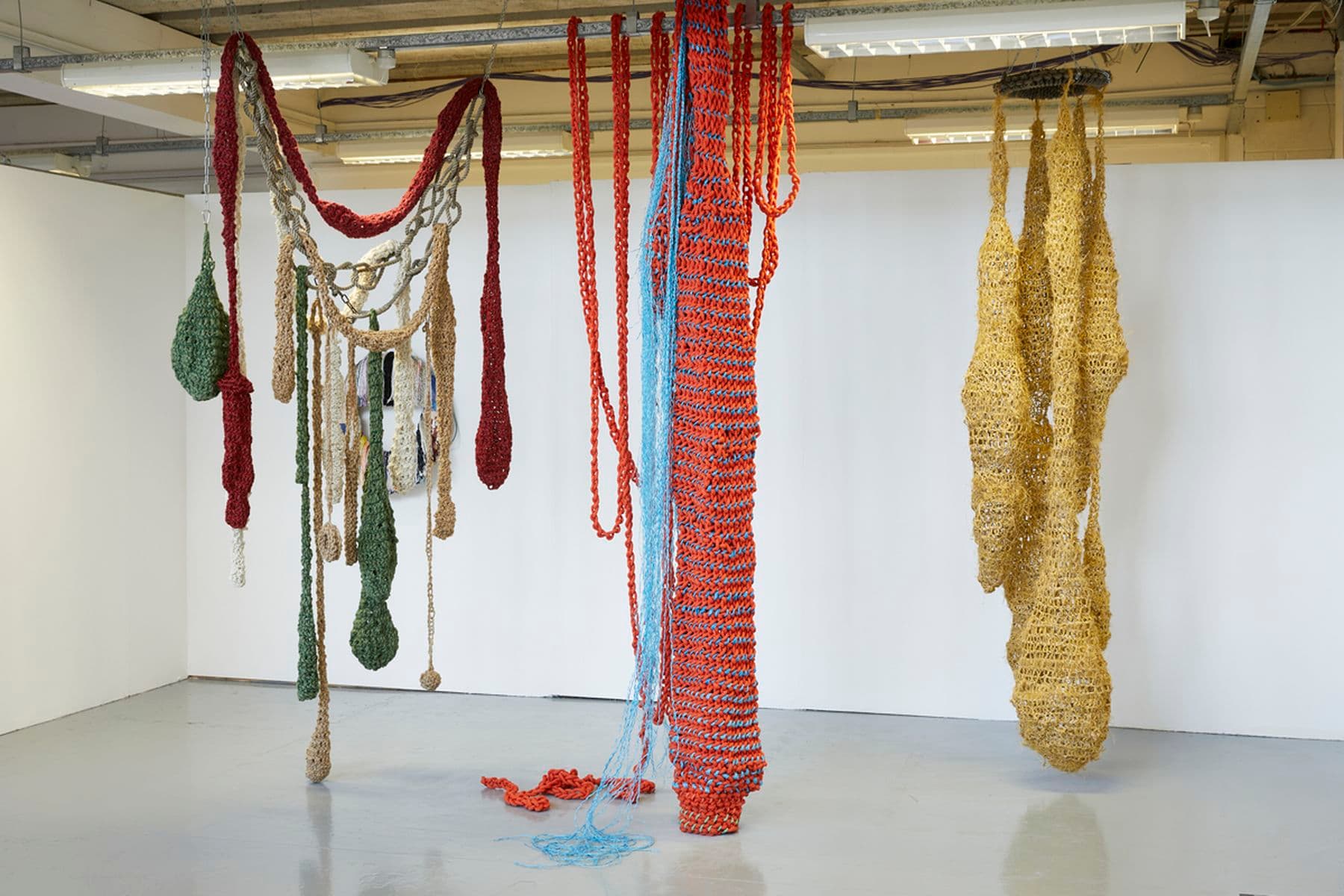 A photograph of three knitted structures hanging from the ceiling. The knitting comprises of greens, oranges, blues, yellows and creams. The structures have varying shapes.