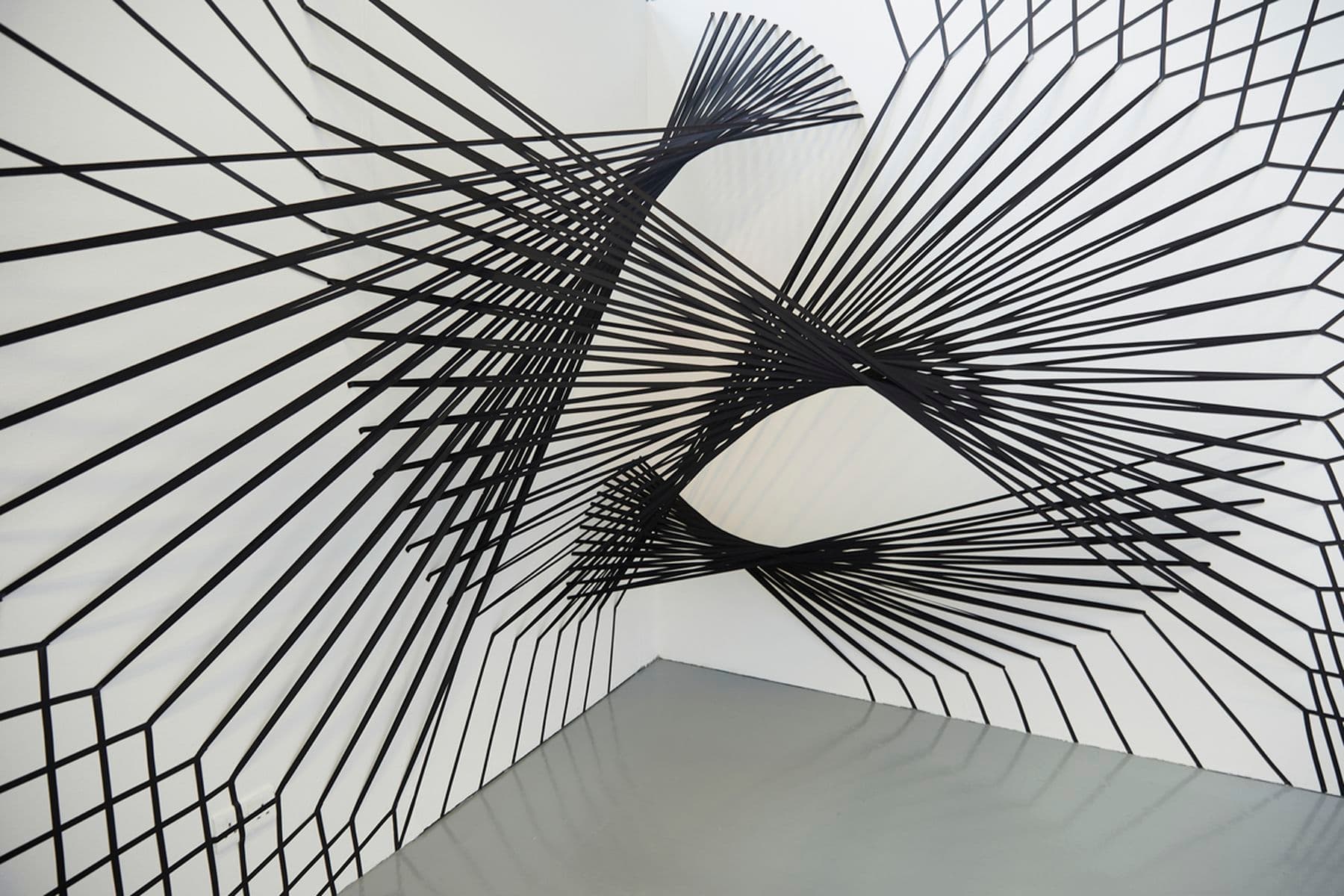 A photograph of black spiral structures mounted against a white wall. There are three interlinking structures in total consisting of vertical lines.