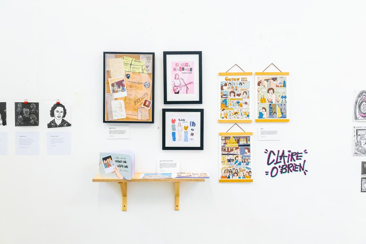 Art exhibition wall featuring framed illustrations, comic strips, and text, with a small shelf holding books.