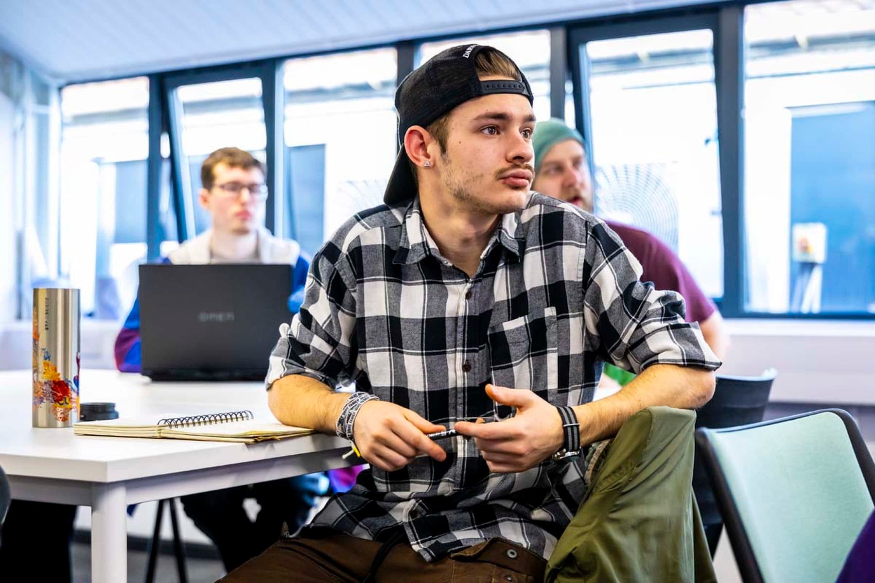 A photograph of three students in a classroom. The student in front is in focus, facing forward, and holding a pencil. The other two are also facing forward, with one using a laptop.