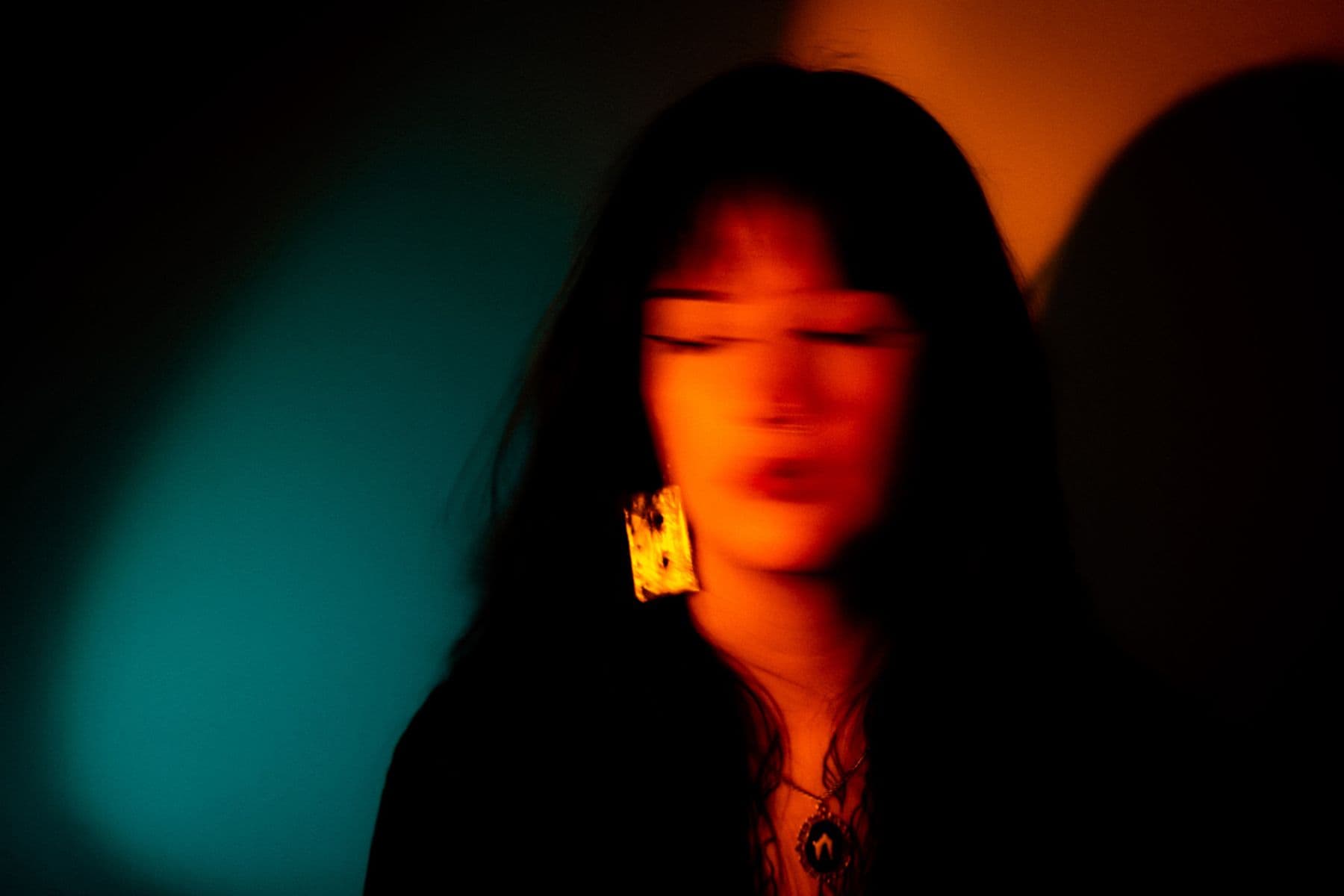 A photograph featuring a person’s face out of focus, they have black hair with a fringe and are wearing chunky gold earrings. The background is half blue and orange with shadows present.