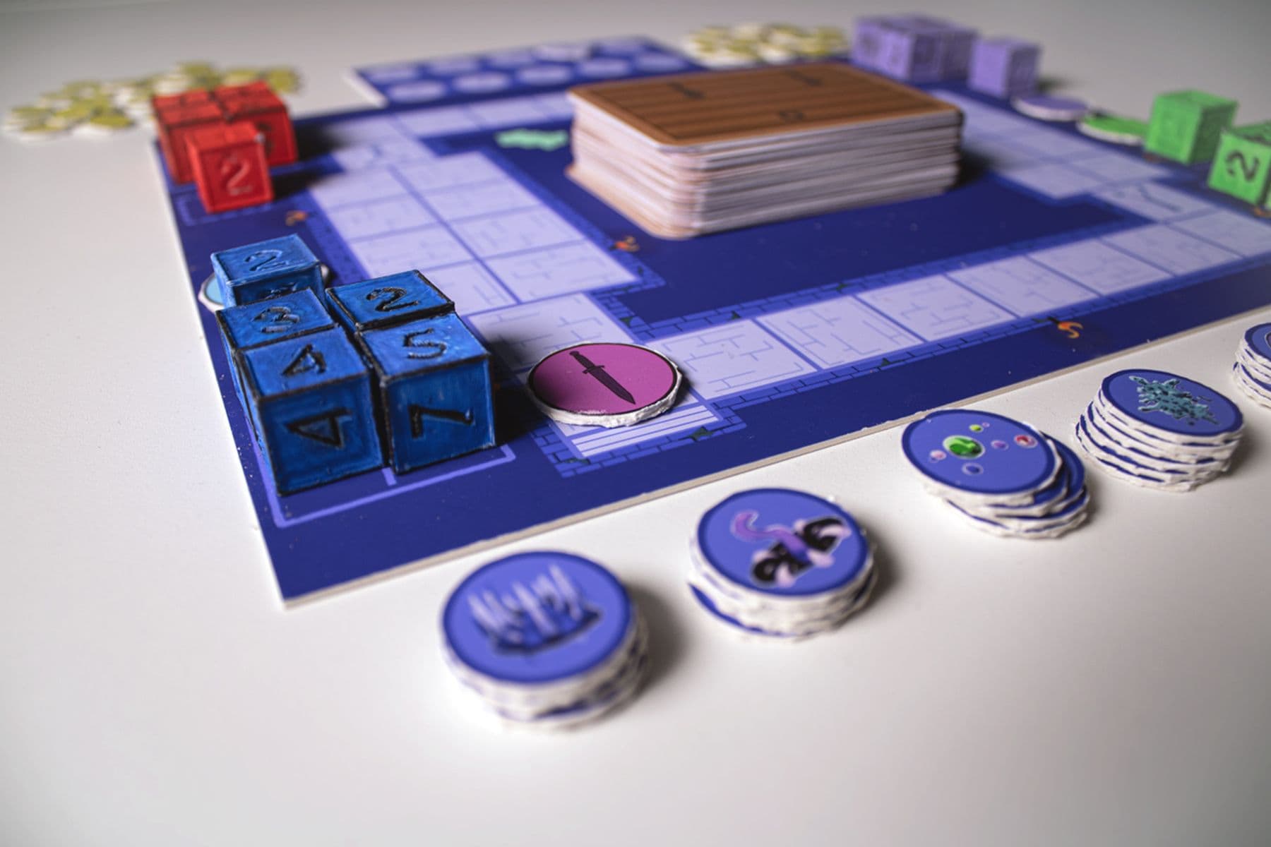 An image of a board game setup with a blue-tiled board, blue dice, stacks of cards and various round tokens with different symbols on top.