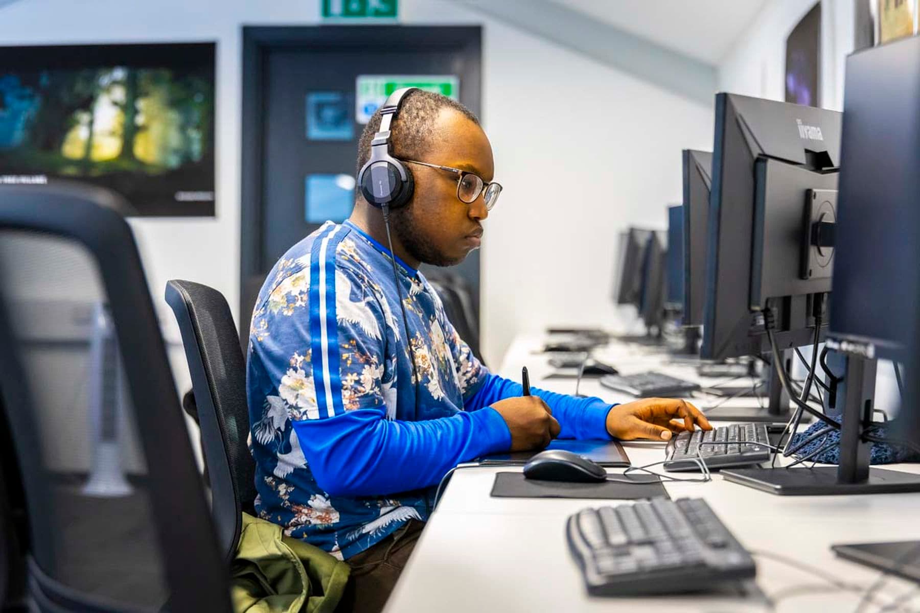 A photograph of a student sitting at a desk, wearing headphones and focused on a computer screen. They are holding a stylus pen and drawing on a tablet in front of them.