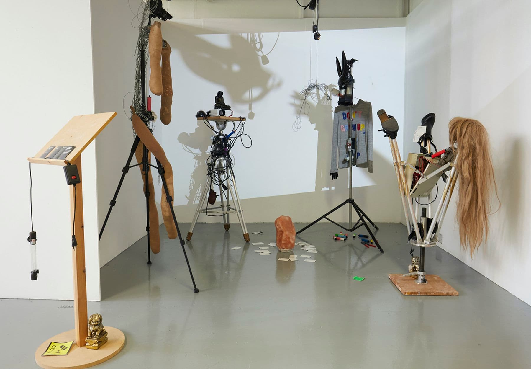 A photograph of an installation featuring a lectern, two tripods and two projector stands, one with a projector on top. They are lined in a row and adorned with different objects including a wig, jumper and small statues. One of the projector stands is upturned. On the floor there are cards, felt tips and crystal.