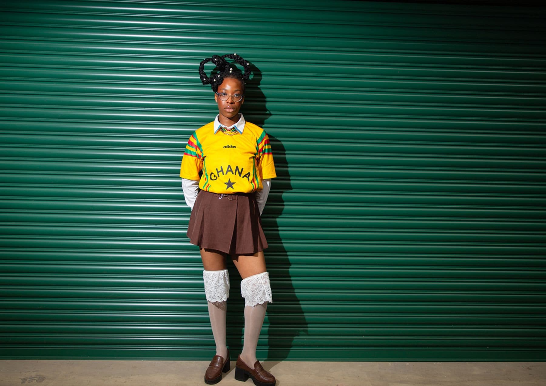 A photograph featuring a person in a Ghana soccer jersey and brown skirt standing against a green corrugated metal wall, wearing knee-high socks and glasses.