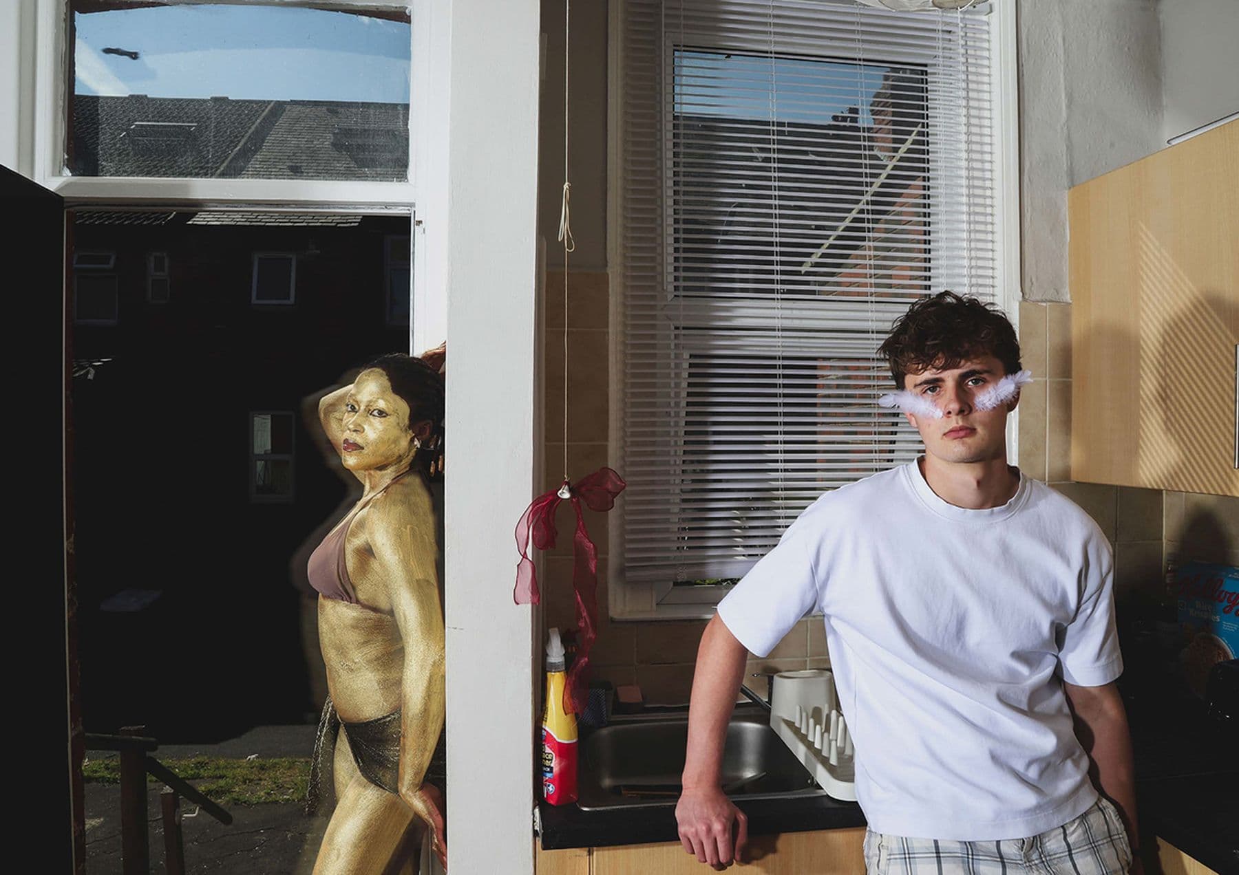 A photograph featuring a person posing in a white t-shirt while standing in a kitchen. In the hallway, a person painted gold and wearing a bikini poses against the wall.