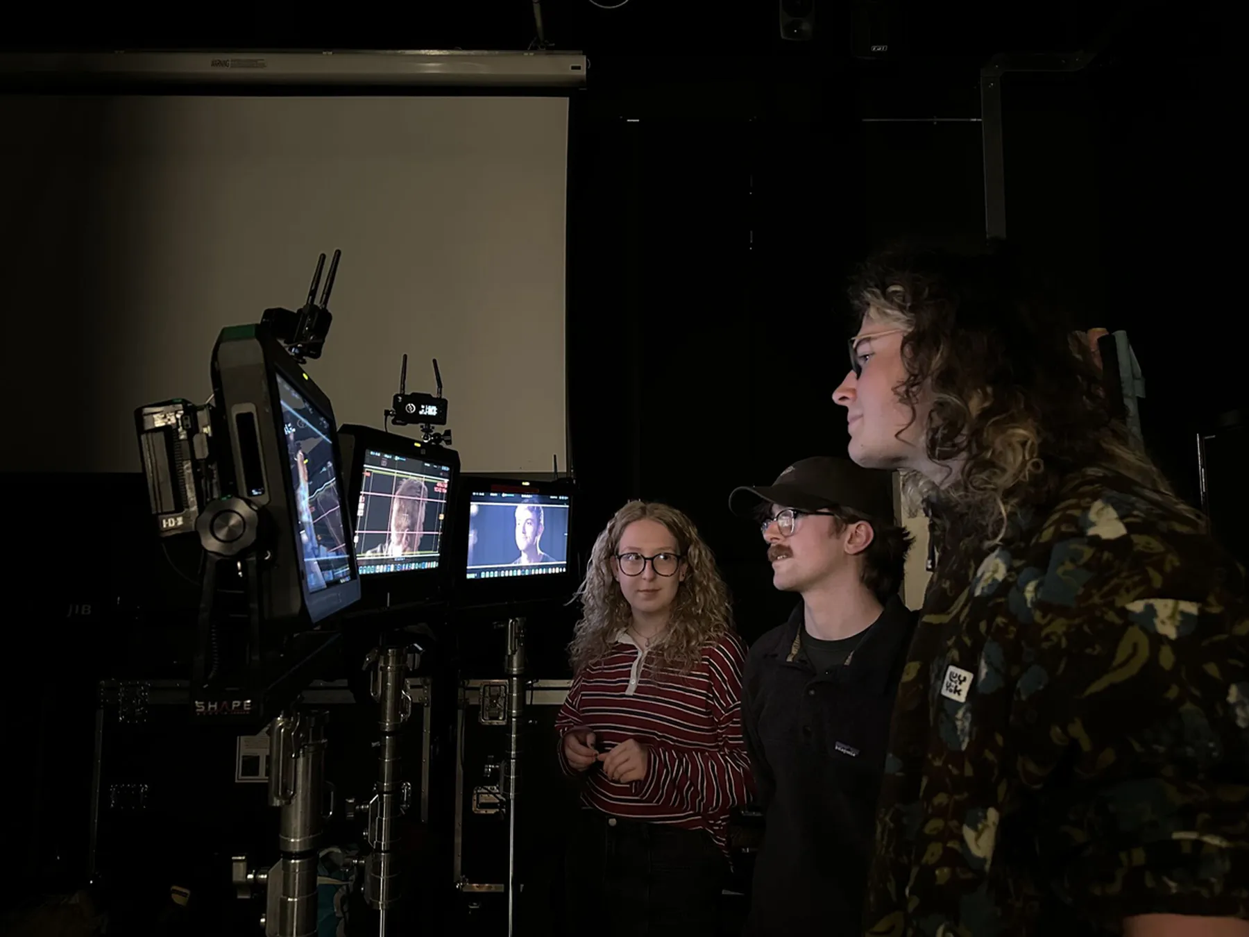 A photograph of three people standing in a dimly lit studio, observing a set of monitors displaying images, in a film or video production setting.