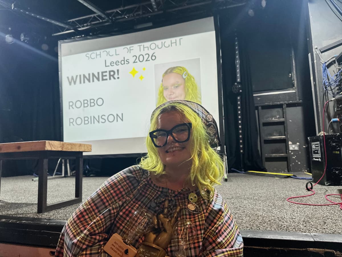Person with bright green hair and large glasses smiling in front of a screen showing "School of Thought Leeds 2026 Winner: Robbo Robinson."