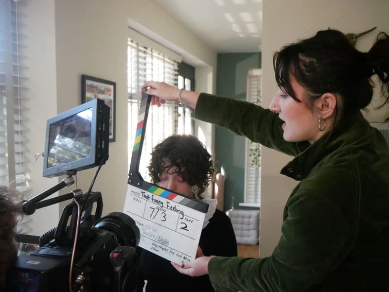 A person holds a film slate in front of a camera, preparing for a scene shoot in a well-lit room.