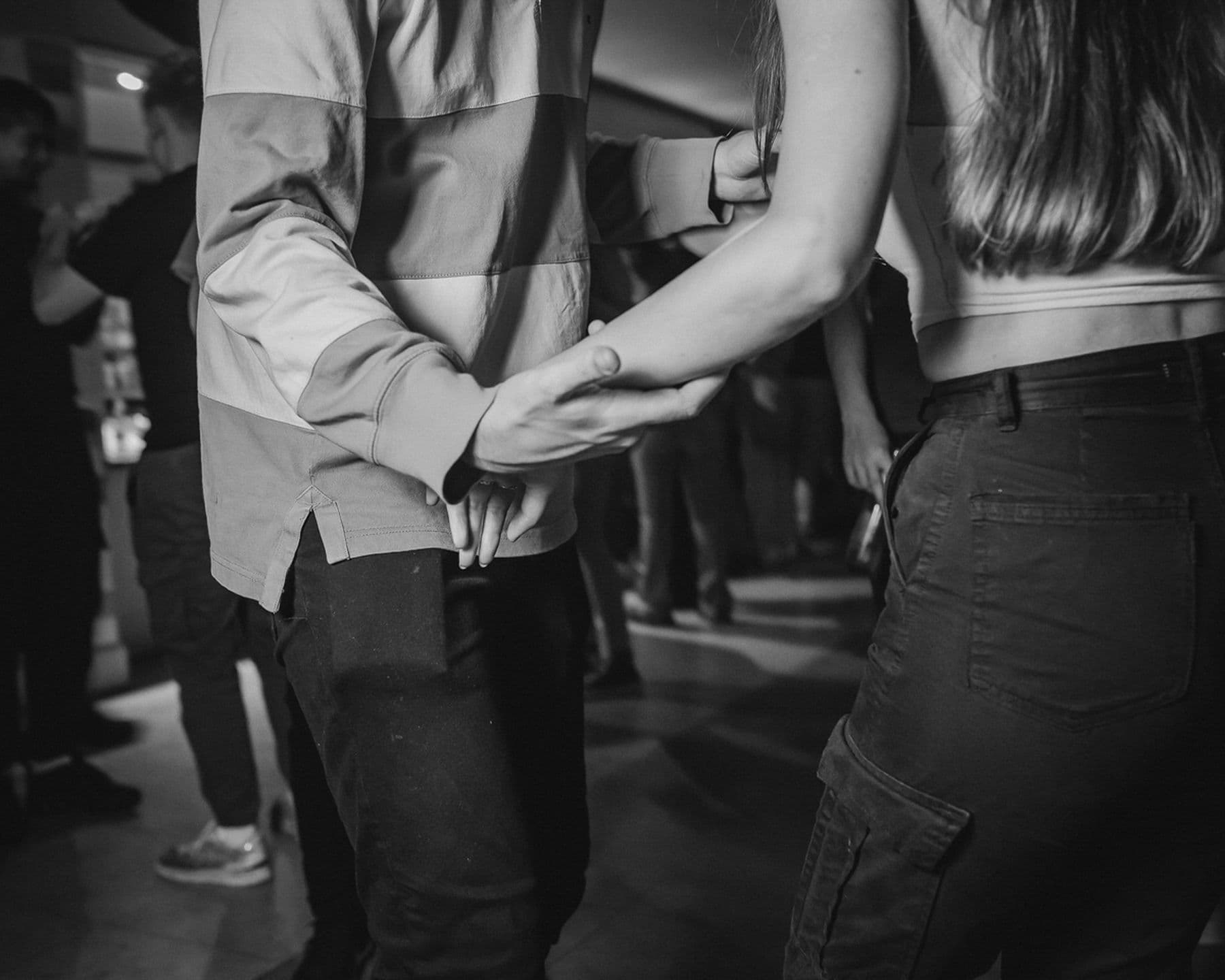 A black and white photograph of two people holding hands and dancing on a dance floor. Only the top half of their bodies and legs are in view.