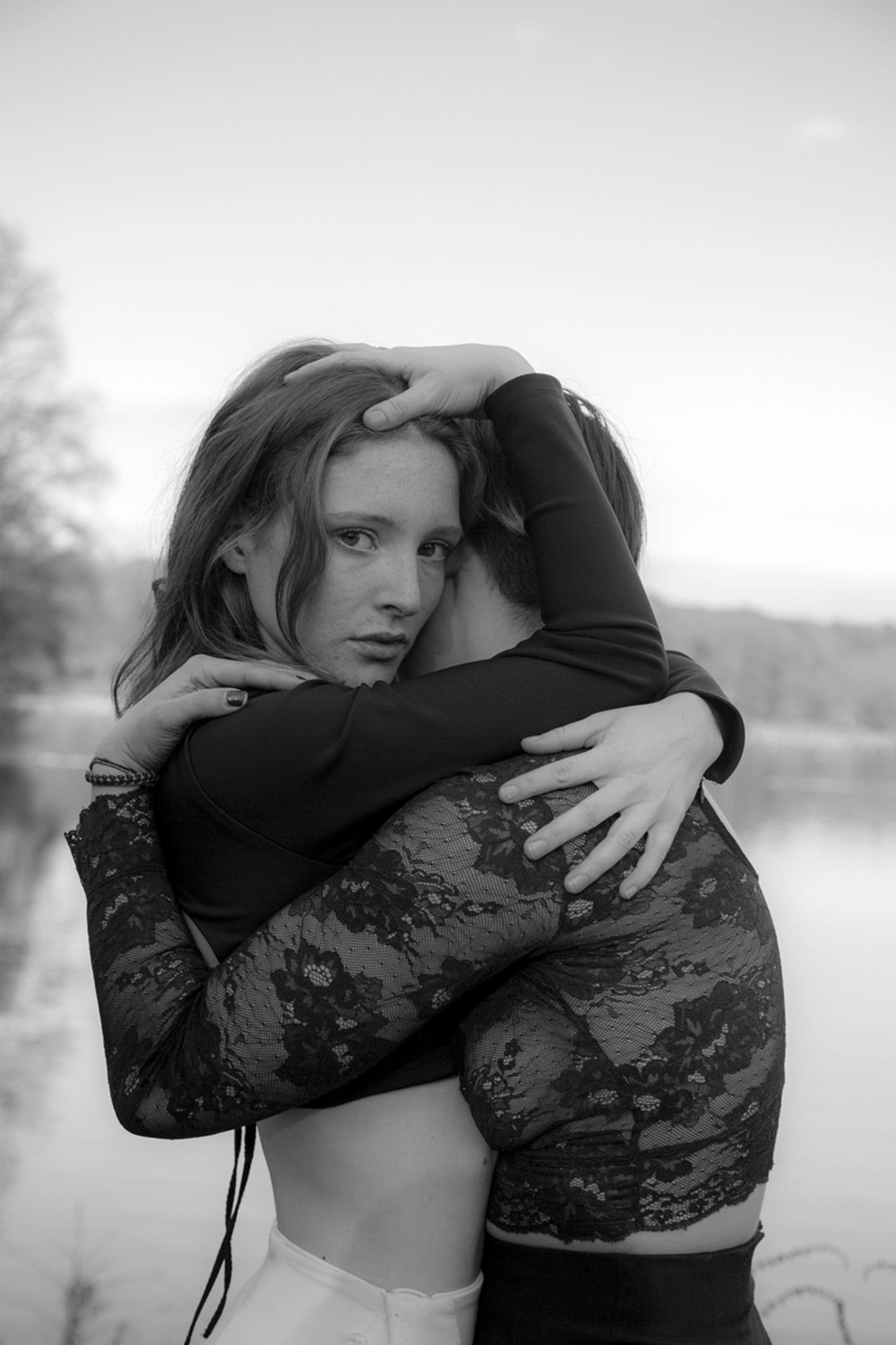 A black and white photograph of two people standing in an embrace. One person is staring directly into the camera, their expression is neutral; the other is facing away and their head is obscured.