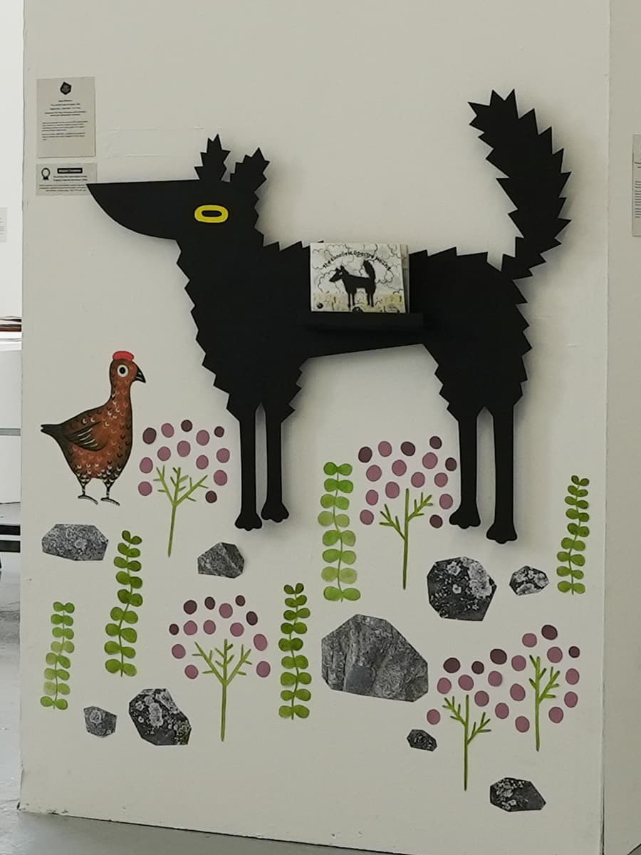 Wall art featuring a black silhouette of a wolf with a small book on a shelf, surrounded by a hen, abstract plants, and stones.