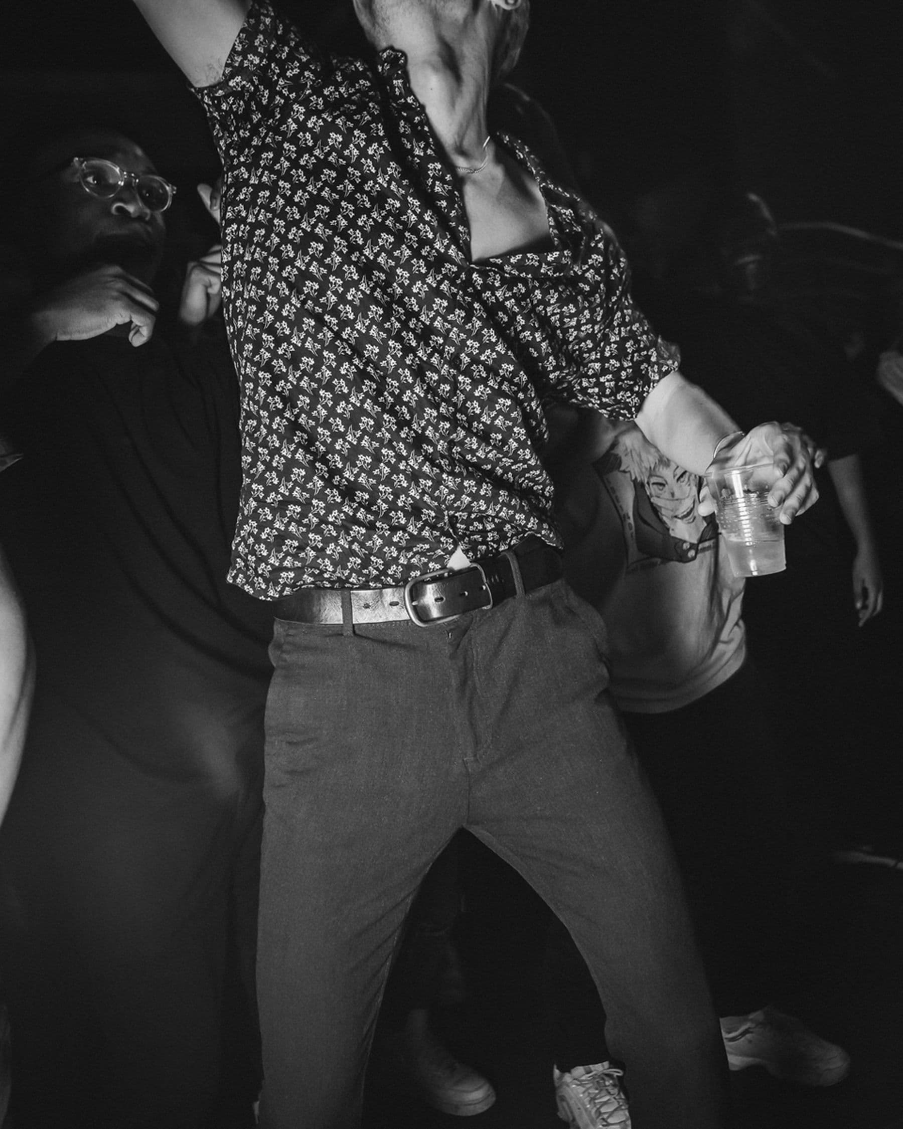 A black and white photograph of a person’s upper body and part of their legs. They are wearing a patterned shirt and holding a plastic cup, one arm is not visible and is likely being raised in the air.