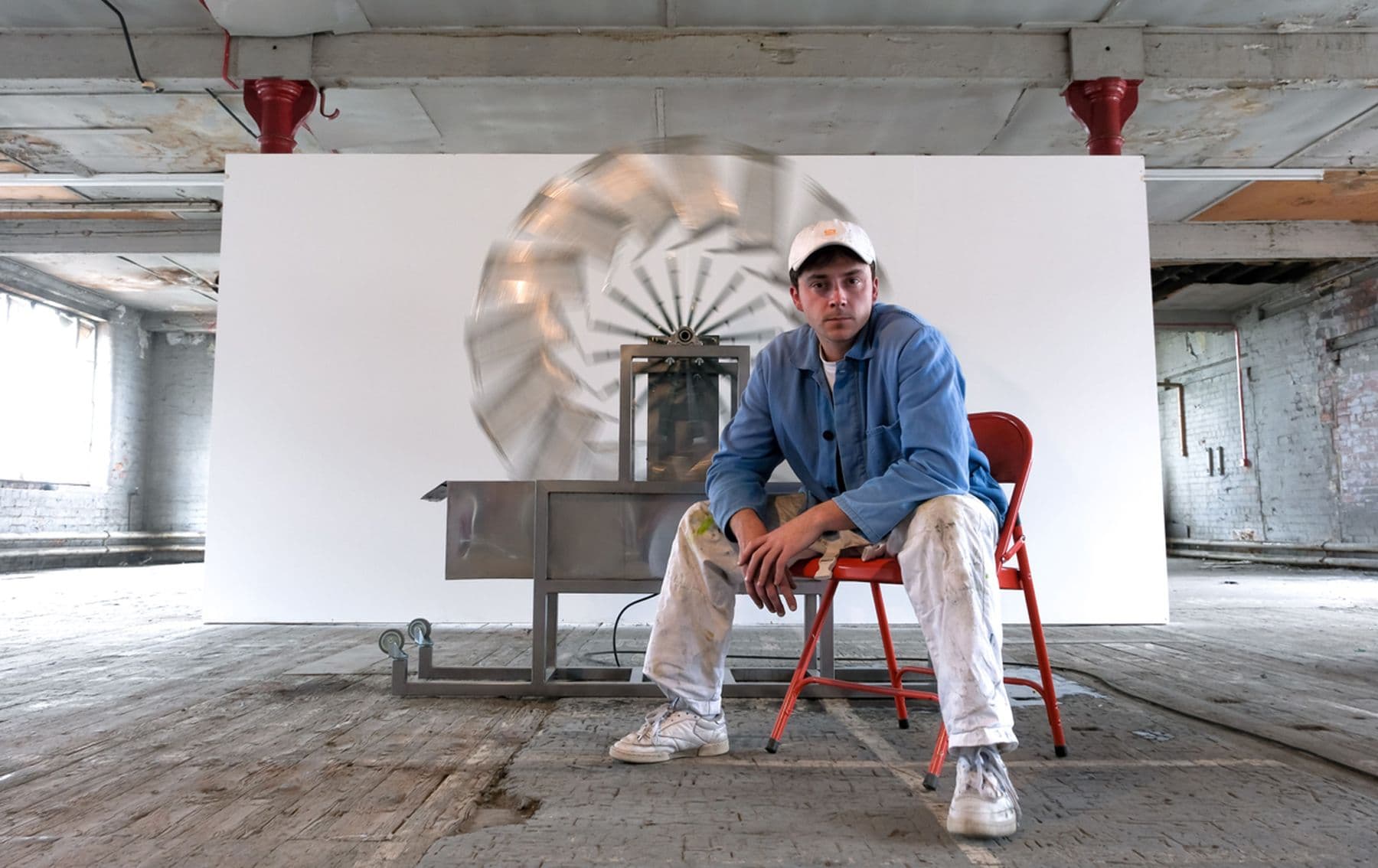 Glen Ogden in a blue jacket and cap sits on a red chair in an industrial loft, paint-stained pants, beside a spinning metal turbine sculpture