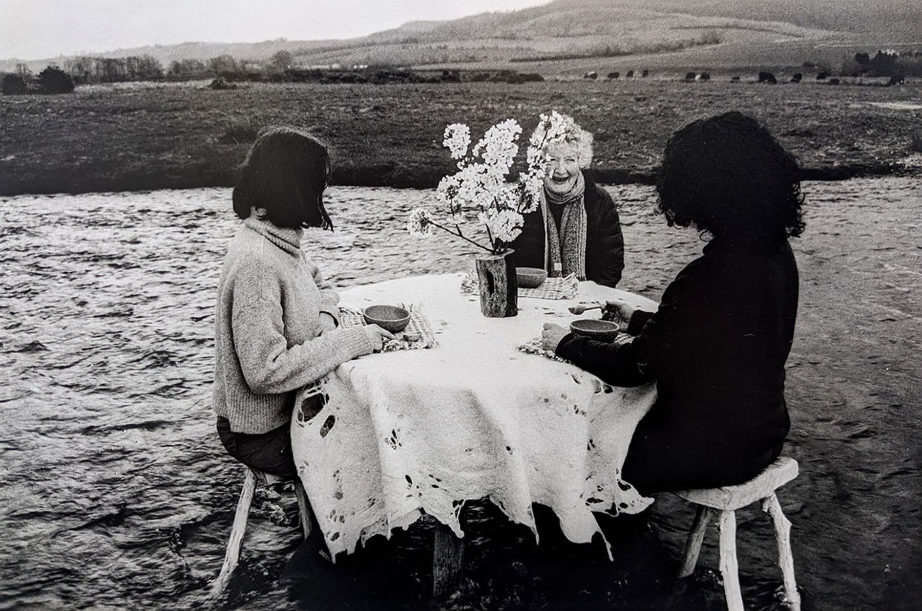 A photograph of three people sitting at a round table with a tablecloth and flowers, dining in the middle of a shallow river, surrounded by a rural landscape.
