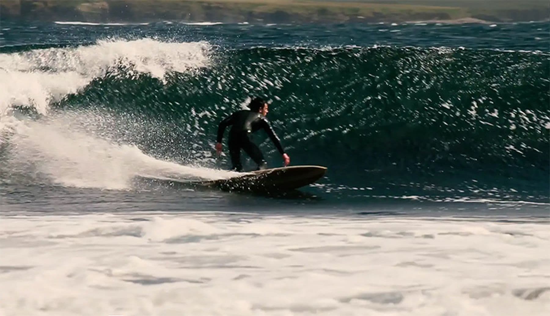 A photograph of a surfer in a wetsuit riding a large wave, with white foam in the foreground and a distant shoreline in the background.