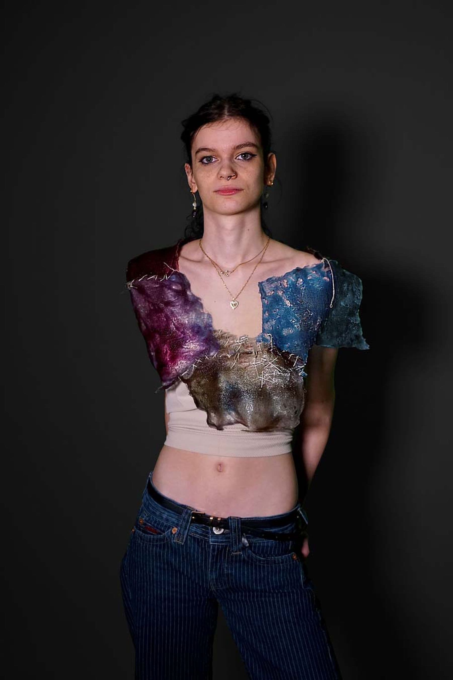 A photo of a person wearing a unique top with colorful, textured fabric pieces whilst standing against a dark background.